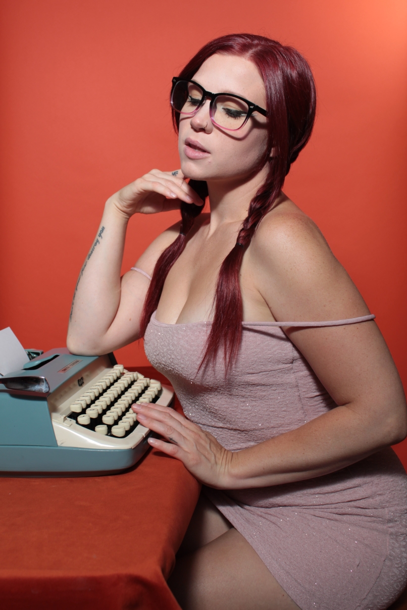 full shot of female sitting with a typewiter. She wears a beige dress against an orange background 