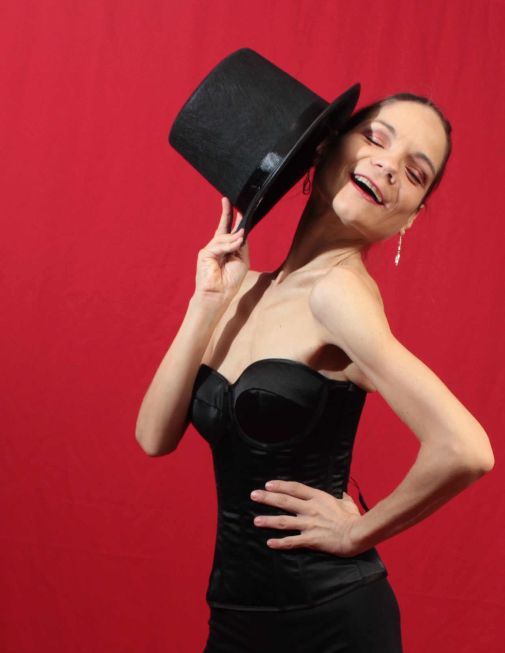 female in black corset, holding a top hat, against a red background